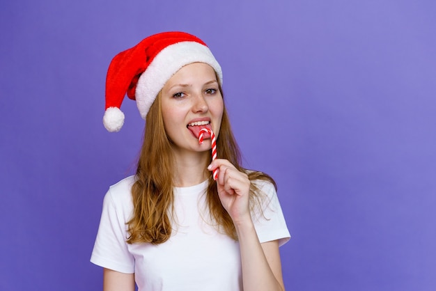 Uma menina alegre com chapéu de Papai Noel está comendo pirulito e rindo em uma camiseta branca mulher jovem e ...