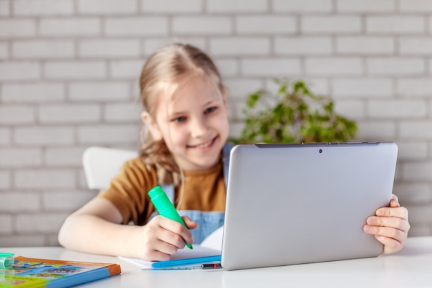 Uma menina alegre realiza trabalhos de casa com um livro e um tablet em casa.