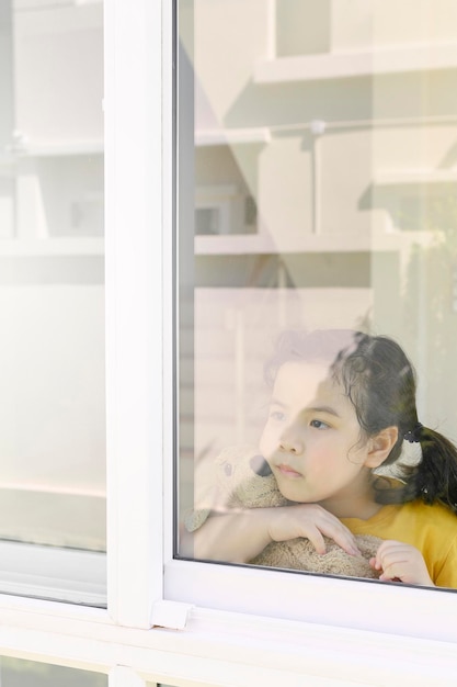 Uma menina asiática fica em casa entediada com o fechamento de escolas devido a uma pandemia macabra. Menina pré-escolar olhando pela janela de sua casa.