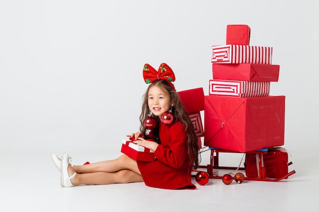 Uma menina asiática sentada em um vestido vermelho senta-se com caixas de presente em um fundo branco. conceito de natal, espaço de texto