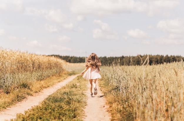 Uma menina atravessa um campo de trigo. Visão traseira. Siga para criança feliz fofa