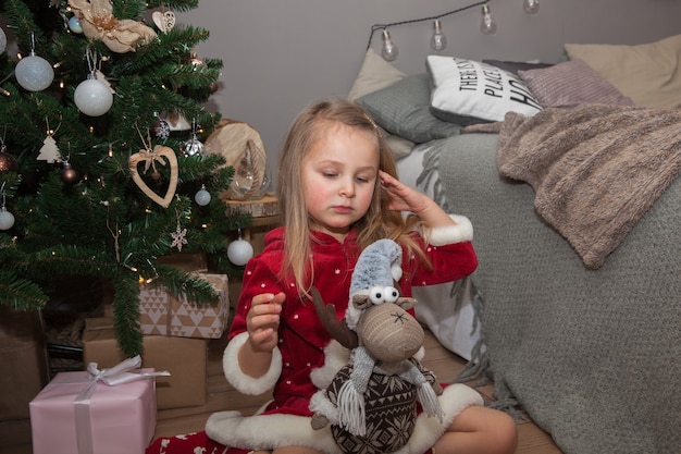 Uma menina bonita com presentes de natal sentada perto da árvore de natal na sala, conceito de ano novo