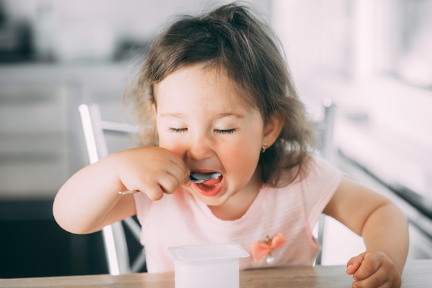 Foto uma menina bonita e engraçada a comer iogurte na cozinha com um vestido rosa à tarde.