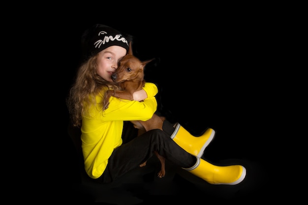 Uma menina bonitinha abraça seu cachorro, o Pinscher Miniatura. Tiro do estúdio em um fundo preto. O conceito de amor pela natureza, proteção dos animais, inocência, diversão. Foto de alta qualidade
