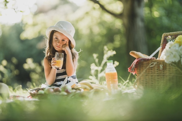 Uma menina bonitinha bebendo suco de laranja e pensando enquanto aproveita o dia do piquenique na natureza.