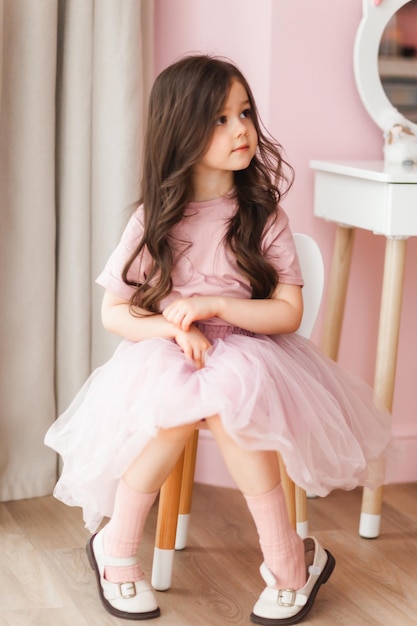 Uma menina bonitinha com cabelo comprido em um vestido inteligente em casa está sentada em uma cadeira em seu quarto