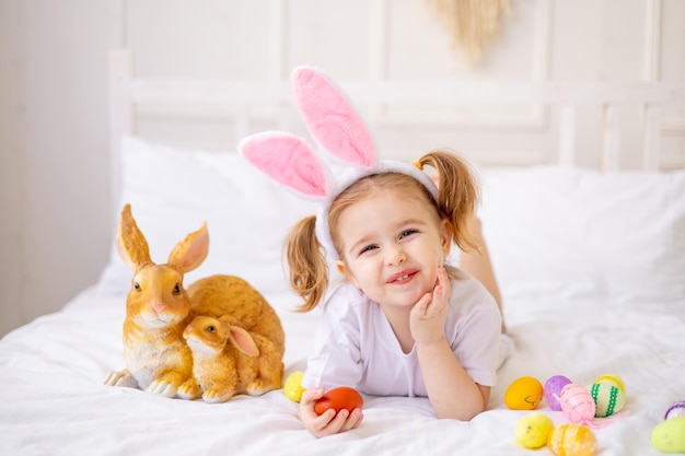 Uma menina bonitinha com orelhas de coelho e ovos coloridos em uma cama branca em casa brincando e sorrindo uma criança loira celebra feliz Páscoa