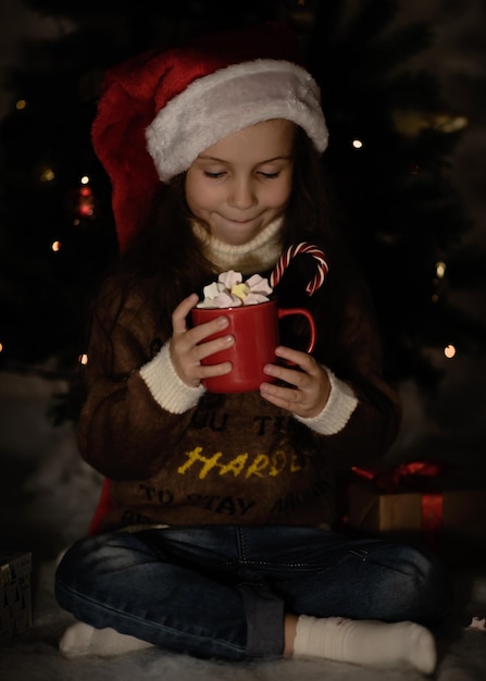 Uma menina bonitinha com um chapéu de Papai Noel com uma caneca de cacau.