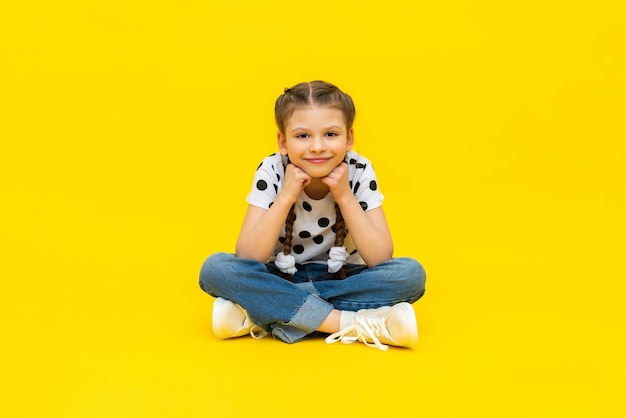 Uma menina bonitinha de jeans azul está sentada sobre um fundo amarelo isolado e sorrindo