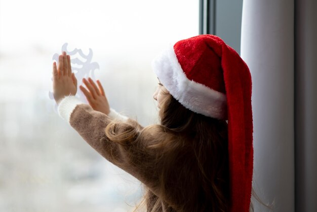 Uma menina bonitinha decora as janelas com flocos de neve Ano Novo Atmosfera de Natal