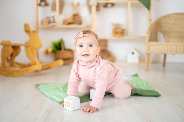 Uma menina bonitinha e saudável de até um ano de idade em um terno rosa feito de tecido natural está sentada em um tapete em um quarto infantil com brinquedos educativos de madeira, olhando para a câmera sorrindo