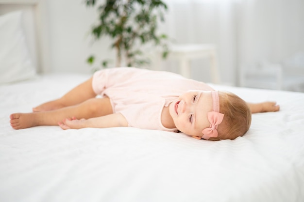 Uma menina bonitinha e saudável de até um ano de idade em uma roupa rosa feita de tecido natural está deitada na cama na roupa de cama branca no quarto olhando para a câmera o bebê está em casa