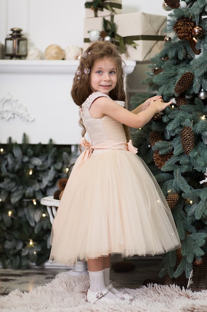 Uma menina bonitinha em um vestido bege exuberante pendura cones na árvore de Natal que está em pé na casa. Conto de Natal, infância feliz.