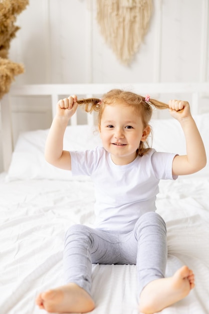 Uma menina bonitinha em uma cama de algodão branco em casa está brincando com rabos e sorrindo a criança está brincando e brincando em casa pela manhã