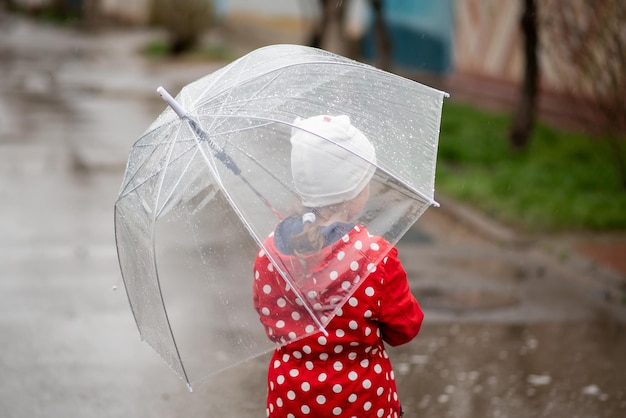 Uma menina bonitinha em uma capa vermelha, botas vermelhas e um chapéu branco pula em poças e se diverte. A garota tem um guarda-chuva transparente nas mãos. Infância feliz. Início da primavera. Emoções.