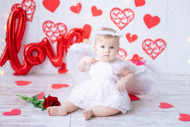 Uma menina bonitinha em uma fantasia de anjo com asas em um fundo de corações vermelhos e a inscrição amam o conceito de dia dos namorados dia dos namorados