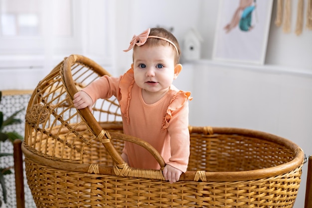 Uma menina bonitinha em uma roupa rosa está de pé em um berço de vime em casa olhando para a câmera sorrindo o sono saudável das crianças