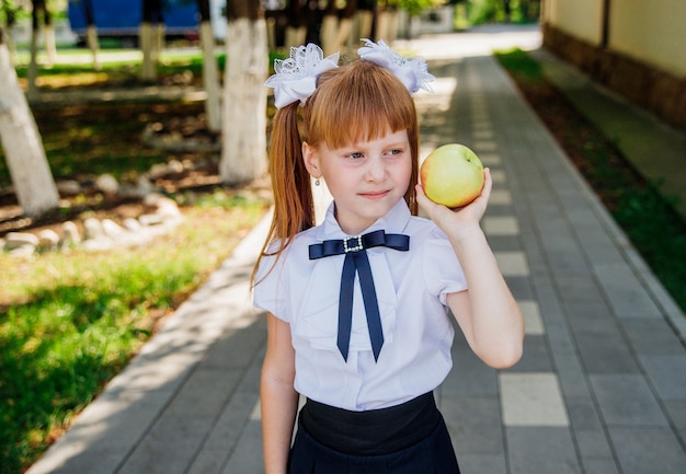 Uma menina bonitinha está de pé no parque ou no pátio da escola e segurando uma maçã verde nas mãos.