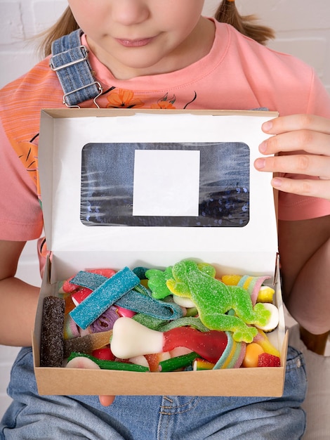 Uma menina bonitinha está segurando uma caixa cheia de doces multicoloridos em suas mãos