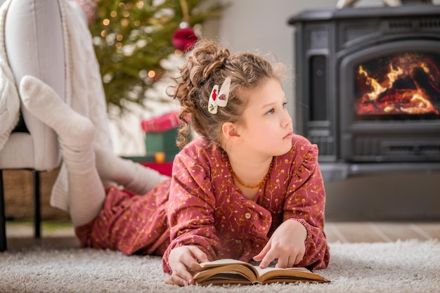 Uma menina bonitinha lê um livro em casa em um interior vivido perto de uma árvore de Natal contra o pano de fundo de uma lareira acesa