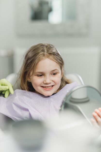 Uma menina bonitinha olhando os dentes no espelho após procedimento odontológico na clínica odontológica.