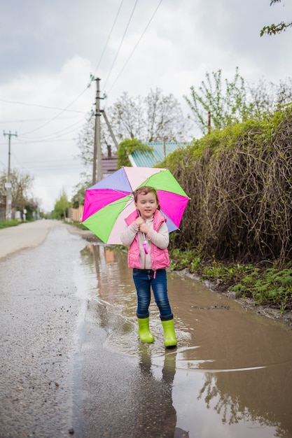 Uma menina caminha através de uma poça grande sob um guarda-chuva multicolorido aberto. Uma linda garota fica em botas de borracha verde e parece.