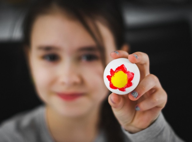 Uma menina caucasiana de colarinho cinza mostra um ovo de Páscoa com uma pintura acrílica amarela