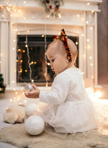 Uma menina caucasiana em um vestido branco fofo e uma bandana com orelhas de tigre está sentada de costas em uma sala perto da lareira com brinquedos para árvores de Natal