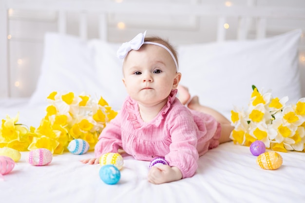 Uma menina caucasiana feliz com uma borda de orelhas de coelho na cabeça está na cama em casa com ovos de Páscoa coloridos e flores amarelas da primavera Olha para a câmera ri bebê de Páscoa