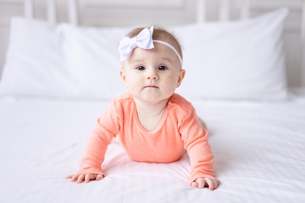 Uma menina caucasiana feliz em uma roupa rosa deitada na cama em casa no quarto na cama branca olha para a câmera ri um bebê saudável