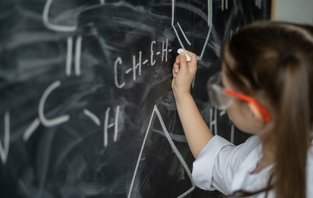 Uma menina colegial perto do quadro-negro resolve um problema de química, jaleco branco e óculos off-line ...