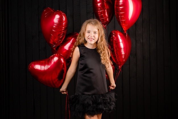 Uma menina com balões em forma de coração manda um beijo em um fundo preto.