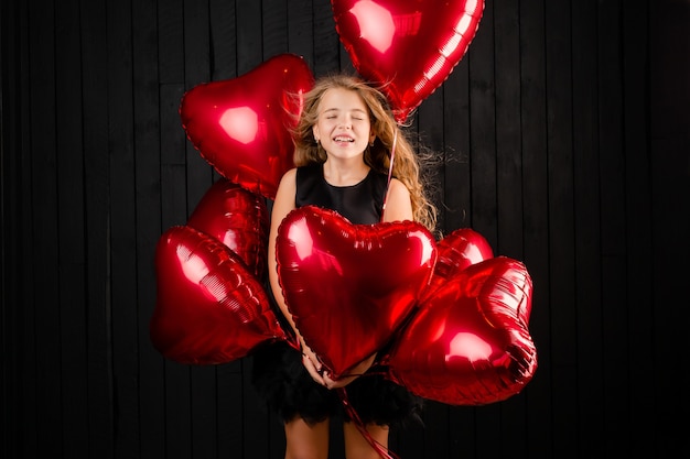 Uma menina com balões em forma de coração manda um beijo em um fundo preto.