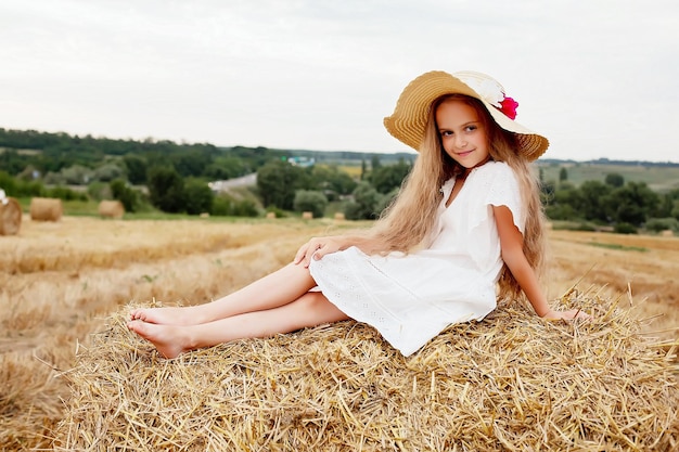 uma menina com cabelo comprido em um vestido branco e um chapéu de palha está sentado em um palheiro