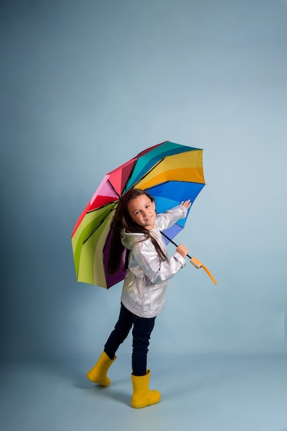 Uma menina com capa de chuva e botas de borracha amarelas segura um guarda-chuva multicolorido sobre um fundo azul com uma cópia do espaço