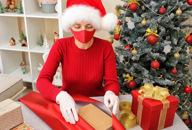 Uma menina com chapéu de Papai Noel, máscara de proteção e luvas está embalando presentes em papel na véspera de Natal