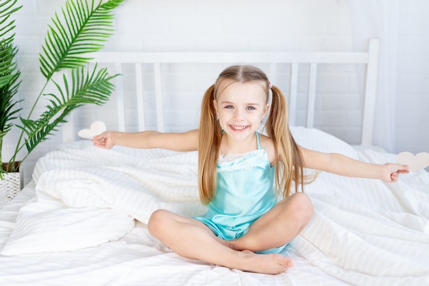 Uma menina com corações nas mãos, sentada em casa na cama em uma cama de algodão branco e sorrindo docemente, conceito de dia dos namorados