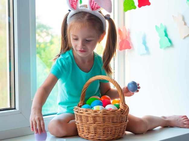 Uma menina com orelhas de coelho no parapeito da janela e sorrindo com uma cesta de ovos de Páscoa.