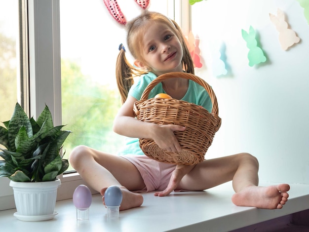 Uma menina com orelhas de coelho no parapeito da janela e sorrindo com uma cesta de ovos de Páscoa.