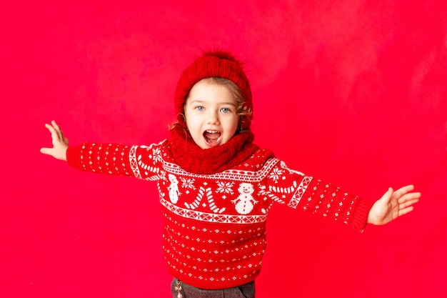 Uma menina com roupas de inverno está feliz, segurando as mãos para o lado em um fundo vermelho. Conceito de ano novo