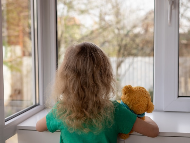 Uma menina com seu ursinho de pelúcia olha pela janela no jardim ficar em casa conceito