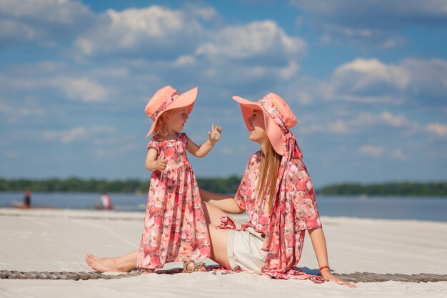 Uma menina com sua mãe combinando lindos vestidos de verão brinca na areia na praia. Aparência elegante de família.