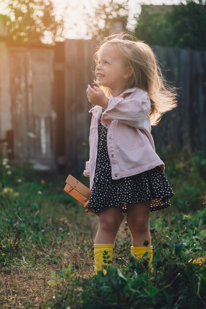 Uma menina com um avião de madeira está em uma clareira ao sol e ri alegremente o riso e a alegria sinceros das crianças feliz infância jogos ao ar livre viajar com um conceito de criança