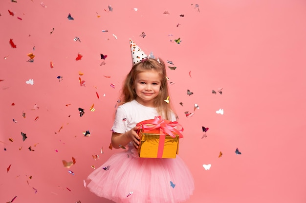 Uma menina com um chapéu de festa segura um presente na frente de um fundo rosa com confete.