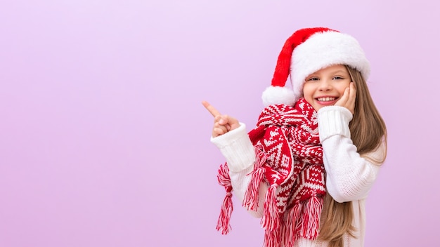 Uma menina com um chapéu de Natal e um cachecol quente de ano novo aponta o dedo para o lado. Fundo isolado.
