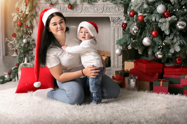 uma menina com um chapéu de Papai Noel abraça seu filho sentado perto da lareira da árvore de Natal