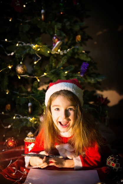 Uma menina com um chapéu de Papai Noel debaixo da árvore de Natal está sonhando