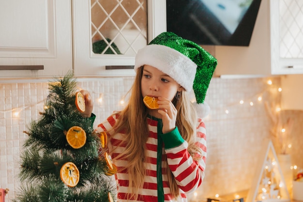 Uma menina com um chapéu de Papai Noel decora a árvore com metades de laranja secas. Um lindo bebê está se preparando para celebrar o Natal.
