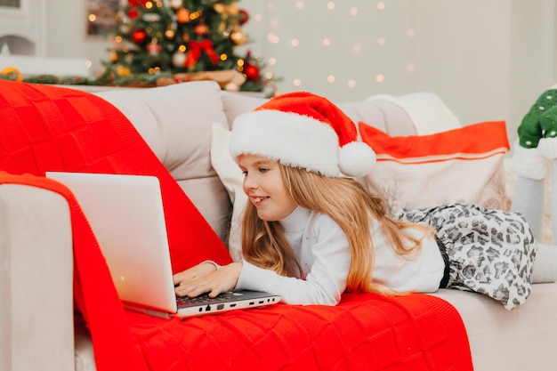 Uma menina com um chapéu de Papai Noel se comunica por videochamada por meio de um laptop, deitada no sofá da sala.