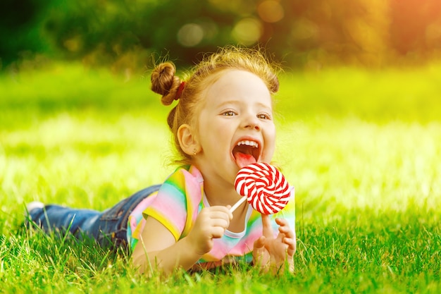 Uma menina com um pirulito vermelho encontra-se na grama de verão no parque.